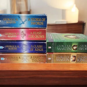 May include: A stack of six paperback books, all by George R.R. Martin, with titles including "A Storm of Swords", "A Feast for Crows", "A Clash of Kings", and "A Game of Thrones".