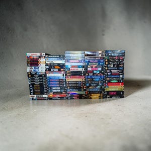 Op de afbeelding: Een grote verzameling rechthoekige VHS-banden gestapeld in vier verticale kolommen. De ruggen van de banden tonen verschillende filmtitels, waaronder Bullitt, Dune, Jaws 2, Independence Day, Dante's Peak, X-Men, Twister en Scarface in kleur.