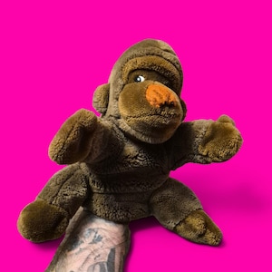 Op de afbeelding: Een bruine pluche gorilla handpop met zachte vacht, een ronde kop, kleine oren en een opvallend oranjegekleurd neusgedeelte. De pop heeft een wijd openstaande houding met de armen gespreid en is ontworpen om op de hand te dragen.