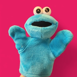 May include: A blue plush Cookie Monster hand puppet with large white eyes and a wide open mouth. The puppet has outstretched arms and is set against a bright pink background. The puppet is made of a soft, fuzzy material.