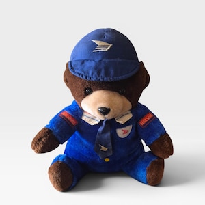 May include: A plush teddy bear dressed in a blue postal worker uniform, complete with a matching cap and tie. The bear has brown fur on its head, paws, and feet. The uniform features the USPS logo and the word "MAIL".