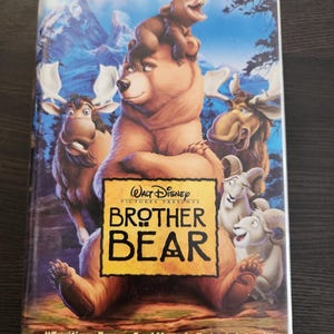 Könnte beinhalten: VHS-Videokassette des Animationsfilms  "Bruder Bär", mit einem braunen Bären mit einem Jungtier auf dem Rücken, umgeben von anderen Tieren, darunter ein Elch und eine Bergziege. Der Titel lautet "WALT DISNEY PICTURES PRESENTS BROTHER BEAR" und enthält ein Zitat aus der Washington Post: "Exciting, Funny And Hugely Entertaining!" - Jane Horwitz, The Washington Post.