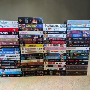 Könnte beinhalten: Eine große Sammlung von VHS-Kassetten, die in vier vertikalen Säulen gestapelt sind. Die Buchrücken zeigen verschiedene Filmtitel wie The Green Mile, Freeway, Footloose, Empire Records, Boogie Nights, Blue Crush und Pearl Harbor mit farbenfrohen Grafiken.