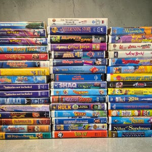 Könnte beinhalten: Eine Sammlung zahlreicher VHS-Kassetten, die in drei vertikalen Säulen gestapelt sind. Die Buchrücken der Kassetten zeigen verschiedene bunte Filmtitel, darunter E.T., The Neverending Story, The Wizard of Oz, Muppets from Space und Stuart Little.