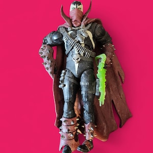 May include: Action figure of Spawn, a comic book character, standing against a pink background. The figure is clad in a black and white suit with a red cape and mask. It holds a green, translucent weapon and has spiked gauntlets and boots.