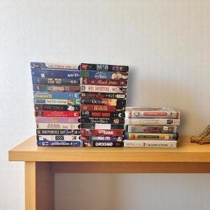 Peut inclure: Plusieurs cassettes VHS empilées sur une table en bois. Les titres incluent "Air Force One", "M.I.B", "Mrs. Doubtfire", "October", "Patriot Games" et "Independence Day". Les cassettes sont de couleurs variées, avec des étiquettes textuelles.