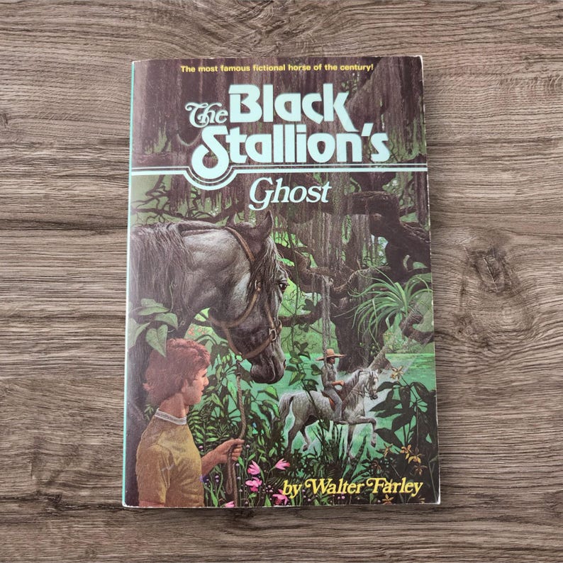May include: A vintage paperback book titled "The Black Stallion's Ghost" by Walter Farley. The cover features a detailed illustration of a gray horse, a young person, and a rider on a white horse in a swampy setting. The book is set against a wooden background.