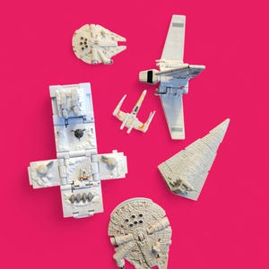 Puede incluir: Una colección de naves espaciales y vehículos de juguete blancos de Star Wars, incluyendo el Halcón Milenario, un X-wing y un Destructor Estelar, sobre un fondo rosa brillante. Los juguetes son detallados y parecen modelos en miniatura.