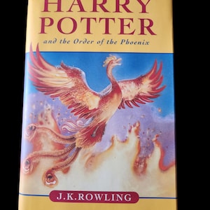 Puede incluir: Un libro de tapa dura titulado "Harry Potter y la Orden del Fénix" de J.K. Rowling. La portada presenta un fénix con plumas rojas y naranjas sobre un fondo azul y morado. El título está en color marrón.