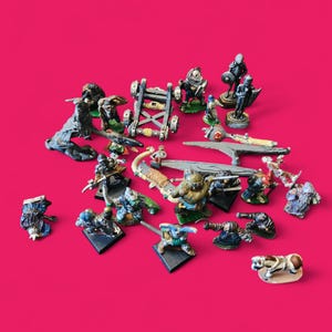 May include: A selection of painted miniature fantasy figures, including warriors and siege engines. The figures are on small bases, with details like armour and weapons. The background is a solid magenta colour.