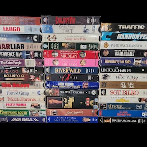 May include: A collection of VHS tapes, stacked on a black background. Titles include "Fargo," "Independence Day," "Forrest Gump," and "Traffic." The tapes are in various colors, with titles in bold, white fonts.