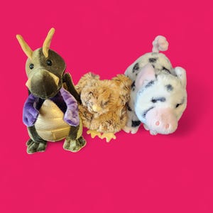 Peut inclure: Trois peluches sur fond rose. Un dragon vert et marron avec des détails violets, un poussin beige et un cochon blanc à taches noires et museau rose. Ces jouets en peluche sont doux et conçus pour le jeu.