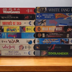 Puede incluir: Una pila de cintas VHS vintage con títulos de películas como "WarGames", "Wayne's World" y "White Fang". Las cintas son de varios colores, con títulos y arte de películas visibles en los lomos. Apiladas sobre una superficie de madera.