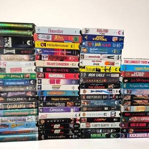May include: A collection of numerous VHS tapes stacked in four vertical columns. The spines of the tapes display various movie titles including Godzilla, Gladiator, The Longest Yard, and Independence Day, featuring diverse colours and graphic designs.