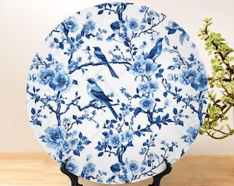 May include: A round decorative plate with a blue and white floral and bird design. The plate features blue birds perched on branches with blue flowers. The plate is displayed on a black stand, showcasing its intricate pattern and color scheme.