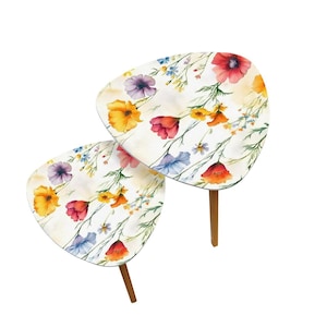 May include: Two nesting tables with a floral design. The tables have a white background with colorful flowers in shades of red, yellow, purple, and blue. The tables have a unique, rounded triangular shape and are supported by three wooden legs.
