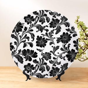 May include: A decorative plate with a black floral pattern on a white background. The plate is displayed on a black stand, showcasing a classic design. The floral pattern includes detailed flowers and leaves.