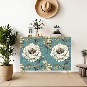 May include: A two-door cabinet with a teal floral pattern featuring large white peony blooms. The cabinet has gold hairpin legs and a light wood top. A straw hat and potted plants are visible in the background.