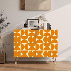 May include: A mid-century modern cabinet with a geometric orange and cream pattern. The cabinet has gold hairpin legs and two doors. Decorative items are on top of the cabinet, including books, a framed picture, and a small black vase.