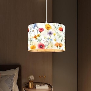 May include: A cylindrical pendant light fixture with a floral pattern. The lampshade features a white background with colorful flowers in shades of red, yellow, orange, and purple. The light hangs from a white cord.