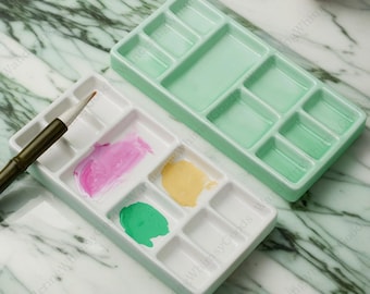 May include: Two ceramic paint palettes, one white with green paint and one green with empty wells. The palettes are made of ceramic and have multiple wells for holding paint.