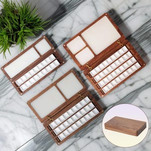 K&ouml;nnte beinhalten: Aquarellpaletten aus Holz mit wei&szlig;en Mischbereichen und zahlreichen kleinen wei&szlig;en Farbn&auml;pfen. Die Paletten sind ge&ouml;ffnet und zeigen das Innendesign. Sie sind aus dunklem Holz gefertigt. Eine geschlossene Palette ist ebenfalls abgebildet.
