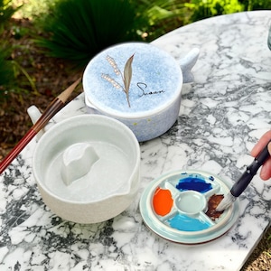 May include: A ceramic watercolour palette with a light blue lid and a matching bowl. The lid features a floral design and the name "Susan". A paintbrush is shown with the palette, which contains various colours.