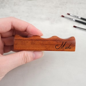 May include: A wooden brush holder with four slots for brushes. The holder is made of reddish-brown wood and has the word "Mate" engraved on the side.