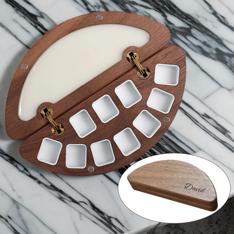 K&ouml;nnte beinhalten: Eine h&ouml;lzerne K&uuml;nstlerpalette mit Scharnierdesign, mit einem wei&szlig;en Harzbereich und mehreren quadratischen Farbn&auml;pfen. Die Palette ist ge&ouml;ffnet und zeigt die N&auml;pfe, und enth&auml;lt eine kleinere, geschlossene Palette mit dem eingravierten Namen "David".