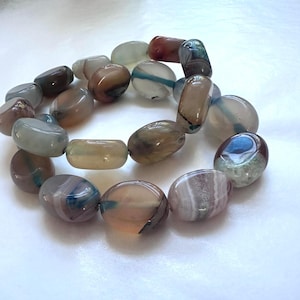 May include: A beaded bracelet featuring polished, oval-shaped stones in shades of brown, gray, blue, and white. The stones are strung together, creating a natural and earthy aesthetic. The bracelet is displayed on a white surface.