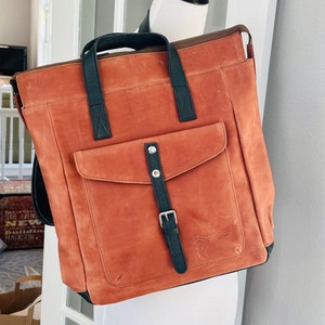 May include: A brown leather backpack with black straps and a buckle. The backpack has a large pocket on the front with a small logo that says "Carry Freedom".