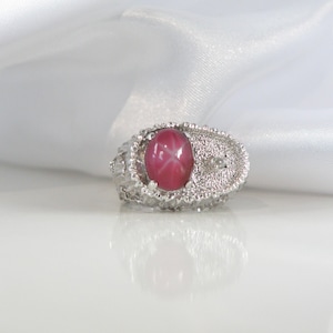May include: A silver ring with a large, oval, pink gemstone displaying a star effect. The ring has a textured band and a small, clear accent stone. The background is a white, silky fabric.