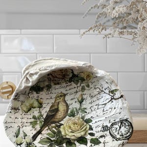 May include: A white ceramic dish shaped like a seashell with a vintage floral design featuring a bird, roses, and handwritten text. The dish has a distressed finish and a wax seal.