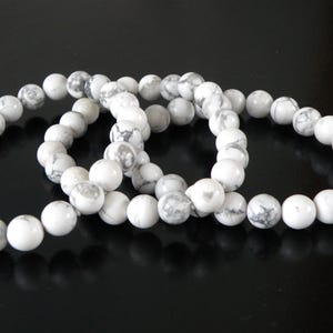 May include: Three white howlite stone bead bracelets. Each bracelet is made of round, polished beads with gray marbling. The bracelets are displayed on a dark, reflective surface.