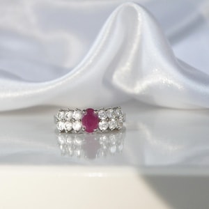 May include: A silver ring with a central oval ruby, flanked by two rows of smaller, clear gemstones. The ring is set against a white, silky background, creating a contrast that highlights the ring's sparkle and design.
