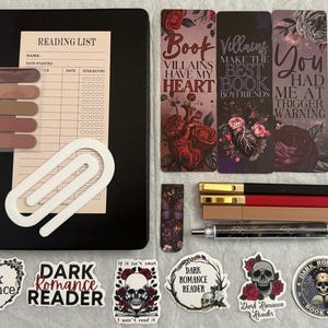 May include: A black notebook with a reading list insert, a white paperclip, five adhesive tabs, three bookmarks with text and floral designs, three pens, one patterned pen, and six stickers featuring skulls and dark romance themes.