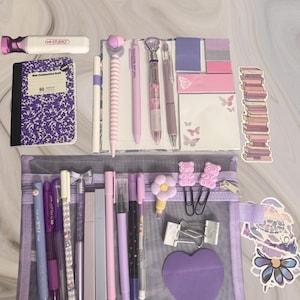 Peut inclure: Une collection de papeterie violette et rose comprenant un mini carnet, divers stylos, des notes adhésives, une trousse en filet, des pinces et des autocollants. Les articles présentent des motifs de papillons, de fleurs et d'ours en nuances de lavande, lilas et rose pâle.