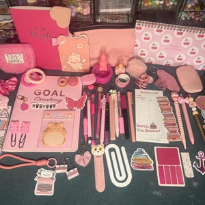 May include: A collection of pink stationery items including a Goal Crushing notebook, a weekly planner with cherry patterns, various pens, stickers, paper clips, a reading list notepad, a fuzzy pouch, and decorative charms in shades of pink and peach.