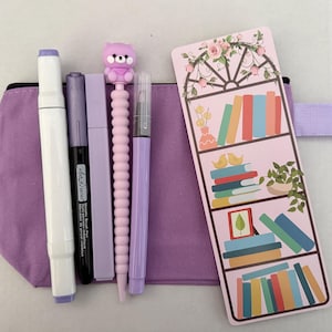 Peut inclure: Une trousse à crayons en toile violette contenant un marqueur blanc, un stylo pinceau métallique noir, un stylo rectangulaire violet clair, un stylo violet clair avec un embout ours et un stylo violet clair, avec un marque-page illustré.