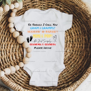 Puede incluir: Body de bebé blanco con texto en varios colores: "So should I call you Gram & Gramps? Mamaw & Papaw? Mimi & Pop? Or Just Simply Grandma & Grandpa? Please Decide." El body está en una cesta tejida.