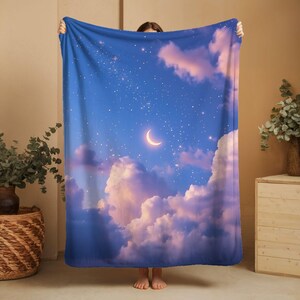 May include: A blue blanket featuring a celestial design with a crescent moon, stars, and fluffy clouds in shades of pink and white. The blanket is held up, showcasing the full design.
