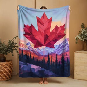 Puede incluir: Una manta rectangular con un gran diseño de hoja de arce roja. El fondo representa un paisaje montañoso en tonos morados, naranjas y azules. La manta se sostiene, mostrando el diseño completo.