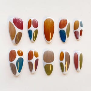 May include: A set of ten almond-shaped press-on nails featuring a white base with various abstract, glossy, multi-colored shapes in shades of red, orange, blue, olive green, and brown. Each nail displays a unique arrangement of these colored segments.
