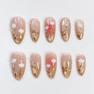 May include: Set of ten oval-shaped false nails. Each nail features a nude base with a gold gradient, adorned with white floral designs, gold vines, and small rhinestones. The nails are arranged on a white surface.