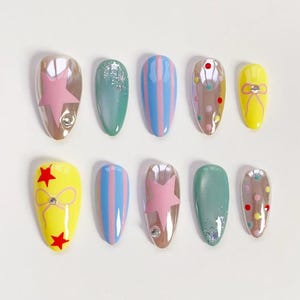 May include: A set of ten artificial nails in various designs. The nails feature colors like pink, blue, yellow, and brown, with patterns including stars, stripes, and polka dots. Some nails have glitter or bow accents.
