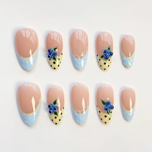 May include: Set of ten oval-shaped false nails. Each nail features a light pink base with a light blue tip. Some nails have a yellow polka dot design with a cluster of blue flower embellishments. The nails are arranged on a white surface.
