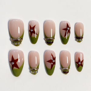 May include: A set of ten press-on nails featuring a nude base with olive green French tips. Some nails are decorated with dark red star accents and small clear rhinestones, while others feature horizontal gold-toned metallic bands.