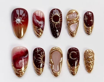 Puede incluir: Un conjunto de diez uñas artificiales con una variedad de diseños. Las uñas presentan una mezcla de colores burdeos, dorados y nude. Algunas uñas tienen adornos dorados, explosiones de estrellas y remolinos. Las uñas son de forma ovalada.
