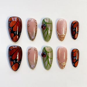 Könnte beinhalten: Ein Set aus zehn mandelförmigen Press-on-Nägeln mit verschiedenen Designs. Zwei Nägel zeigen orange-schwarze Schmetterlingsflügelmuster, zwei sind nude mit goldenen Schnörkeln, zwei sind salbeigrün mit Blattadern und einem 3D-Marienkäfer, zwei sind nude mit goldenen Glitzerspitzen und zwei sind kleine Schmetterlingsflügel-Akzente.