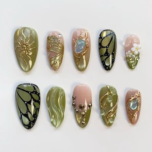 May include: A collection of ten artificial nails in various designs. The nails feature a range of colors, including olive green, pink, and gold. Some nails have intricate gold embellishments, while others showcase butterfly and floral patterns.